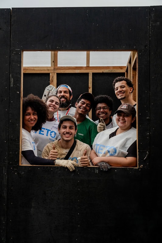 Grupo de voluntários e moradores sorri dentro da estrutura de uma casa em construção no Jardim Lapenna, zona leste de SP.