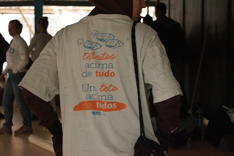 Pessoa com camiseta da TETO Brasil durante mutirão de moradia em comunidade; frase na estampa reforça o compromisso com moradia digna como direito básico.