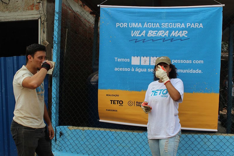 Dois voluntários tomam água em frente a cartaz sobre projeto Água Segura, na Vila Beira Mar (RJ)
