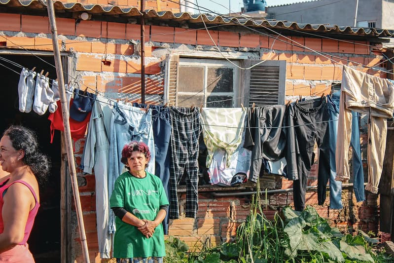 Senhora de cabelos curtos e escuros com camiseta verde em frente à casa em comunidade impactada pela TETO