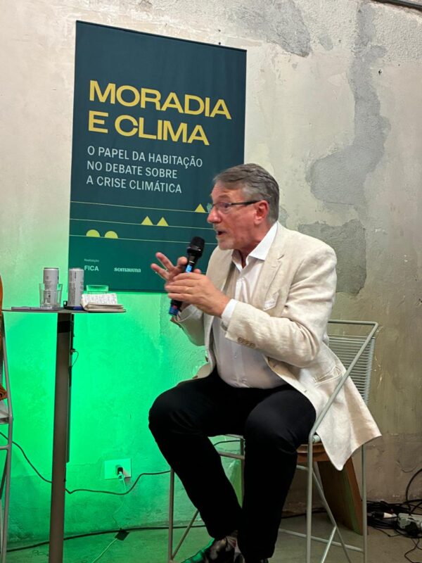 Eduardo Trani, debatedor da mesa de Habitação emergencial em áreas informais e de risco, segura microfone e aparece sentado em frente a cartaz escrito ''Moradia e clima''