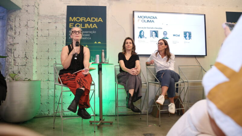 Debatedoras da mesa Locação Social e Gestão aparecem sentadas em frente a painel e tela de projeção com o título do evento sobre crise climática e moradia.
