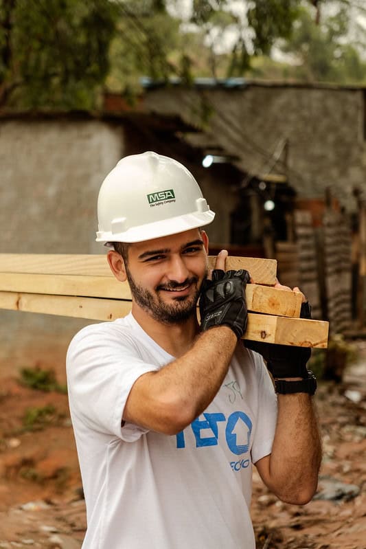 Voluntário usa capacete branco e carrega madeira no ombro