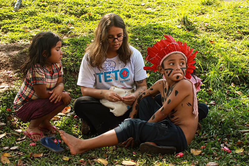Voluntária da TETO sentada na grama no meio de duas crianças