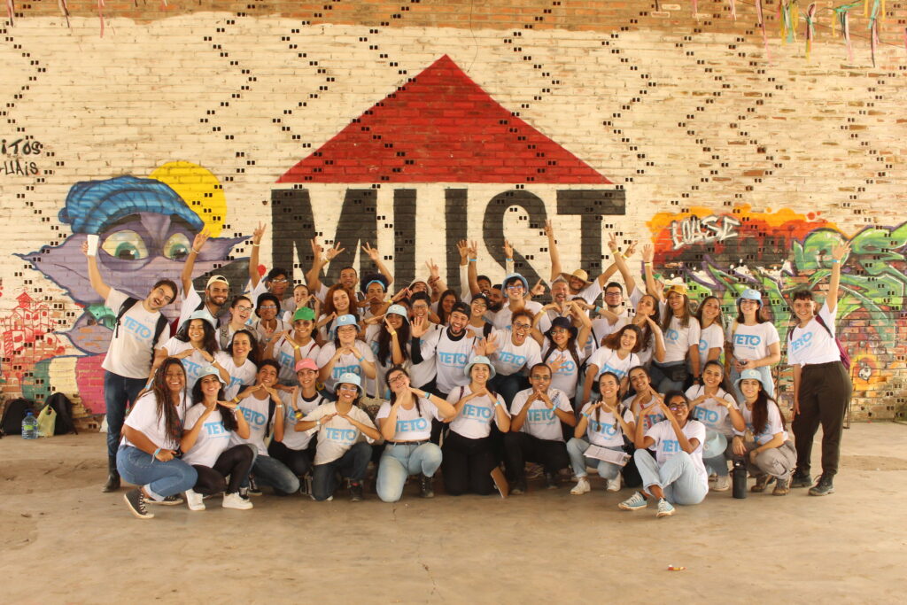Voluntários da TETO posam em frente à parede colorida com desenho e sigla ''MUST''