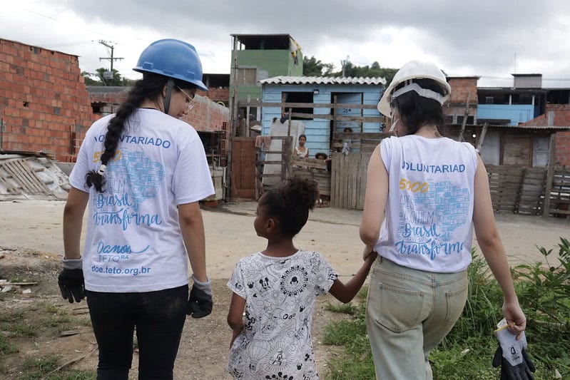 Duas voluntárias da TETO, de costas, levam menina moradora da comunidade pela mão.