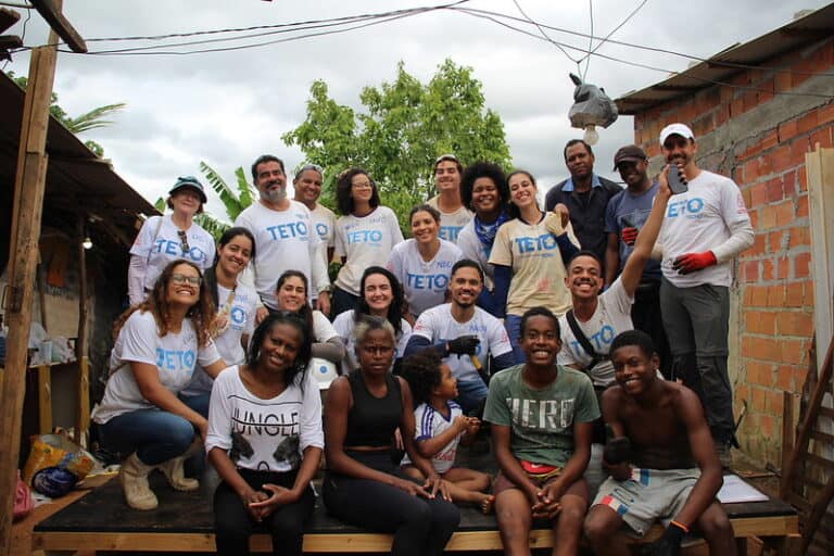 Voluntários e moradores sorriem juntos durante o processo construtivo. Imagem transmite união, orgulho e espírito coletivo após a finalização de uma das 20 moradias resilientes construídas em parceria com o Airbnb.