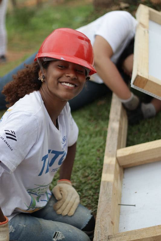 Voluntária da TETO, com capacete vermelho e luvas, sorri enquanto constrói uma das casas resilientes. A madeira e os parafusos à sua frente evidenciam o processo de construção coletiva.