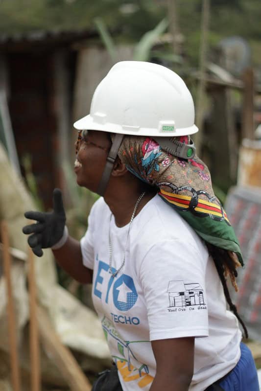 Voluntária da TETO, sorridente, com capacete branco e luvas de construção, durante construção em comunidade.