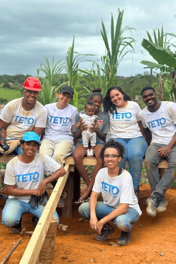 Voluntários sorridentes ao lado da moradora e sua bebê em uma área de construção com chão de terra vermelha e vegetação ao fundo. Todos vestem camisetas da organização, alguns com capacetes e luvas.