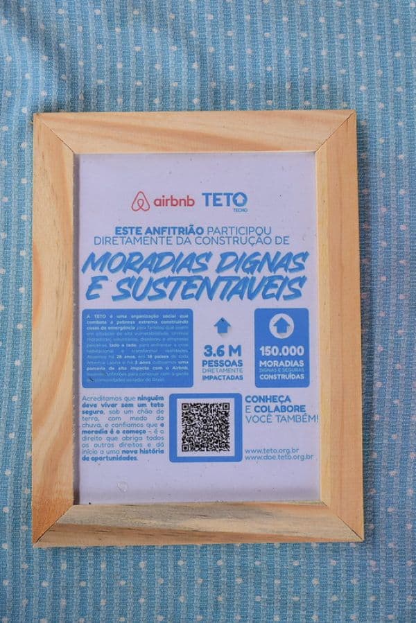 Quadro de madeira com certificado do Airbnb e da TETO sobre participação de anfitriões na construção de moradias resilientes. O texto destaca o impacto social da organização, com 3,6 milhões de pessoas diretamente beneficiadas e 150 mil moradias construídas.