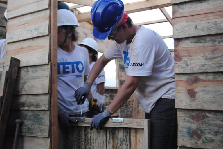Voluntário da TETO, com capacete azul e luvas, instala dobradiça em porta durante mutirão de construção. Ao fundo, outros voluntários trabalham em equipe, com camisetas da organização.