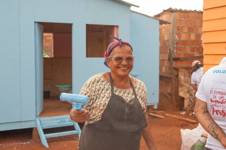 Moradora sorridente segura um rolo de tinta azul em frente à casa recém-construída. Ela veste avental, lenço na cabeça e óculos, representando o protagonismo comunitário na construção da própria moradia.
