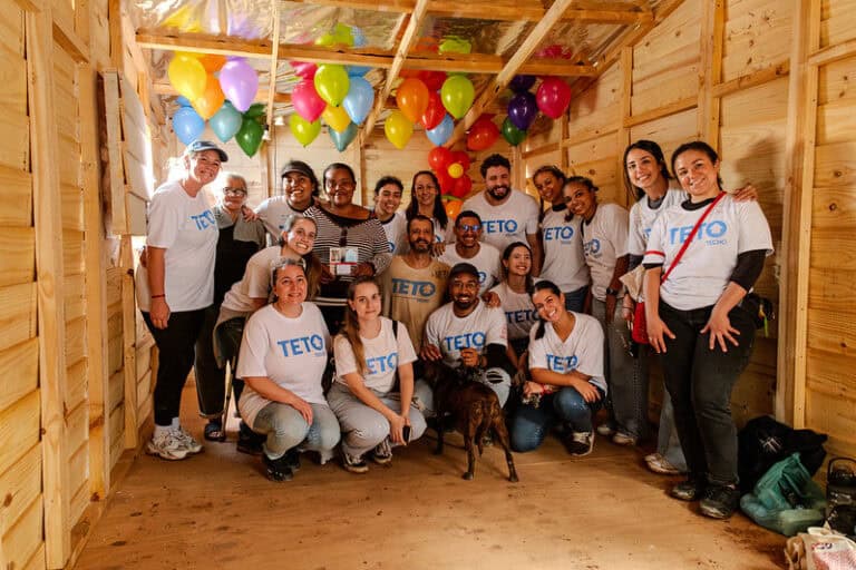 Moradora com voluntários da TETO Brasil durante entrega de moradia digna, celebrada com balões coloridos e emoção.