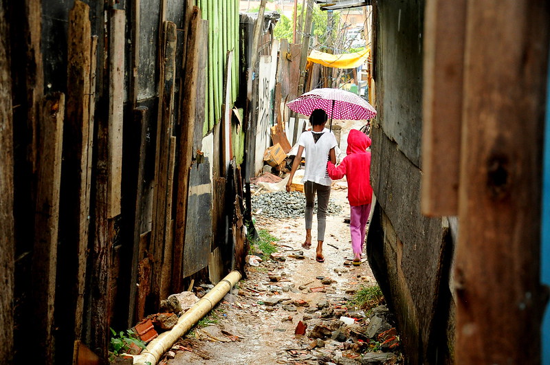 Duas crianças caminham por um beco de favela sob chuva, compartilhando um guarda-chuva.