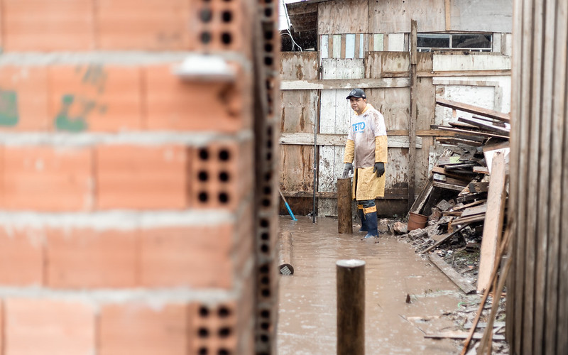 Voluntário da TETO Brasil com galochas caminha por área alagada na Vila União, na periferia de Curitiba (PR), evidenciando as condições de vulnerabilidade climática enfrentadas por famílias em assentamentos informais e o impacto da falta de moradia adequada no cotidiano das comunidades.