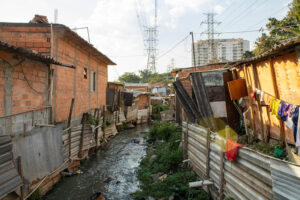 Moradias da Favela City Jaraguá, em São Paulo (SP), às margens de um córrego, onde tubulações despejam esgoto a céu aberto, evidenciando a precariedade de saneamento e os riscos à saúde no território.