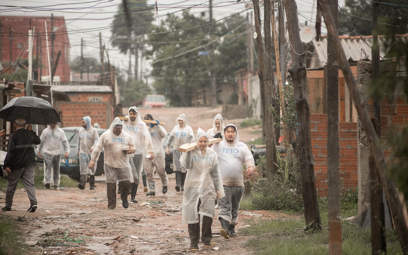 Voluntariado da TETO Brasil carregam madeira para construção de moradia emergencial sob chuva no Paraná.