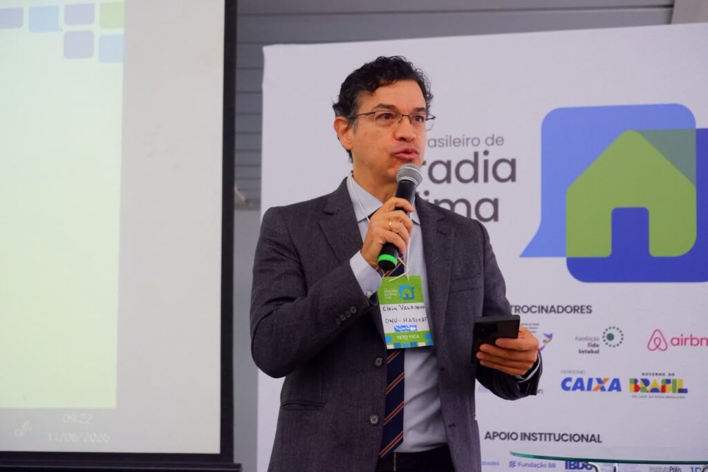 Apresentador de óculos segura microfone durante Fórum de Moradia e Clima.